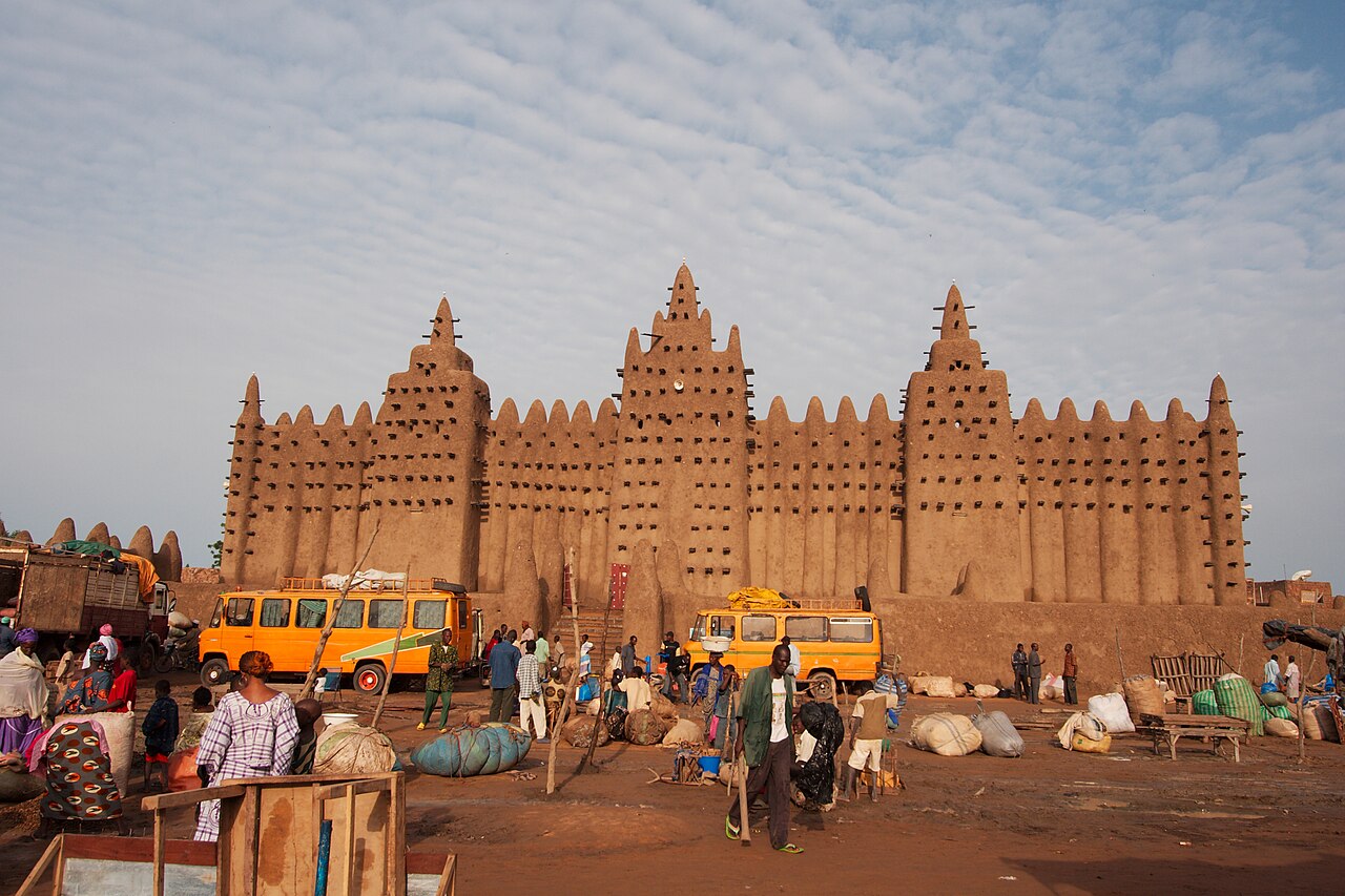 The Great Mosque of Djenné, Mali — the largest mud-brick (adobe) building in the world, rebuilt in 1907 on a 13th-century foundation, its three towers and forest of wooden armatures (torons) rising above the Monday market flood plain of the Bani River, with the town of Djenné spread across the island around it