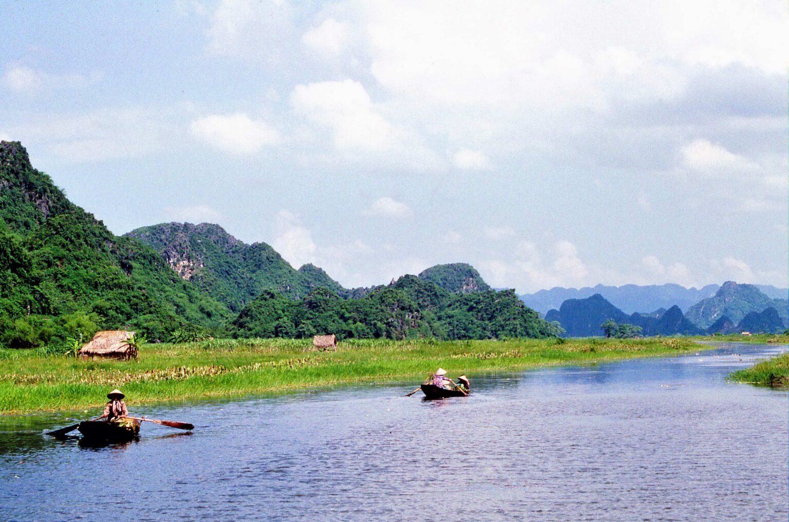 Forested limestone towers rising straight out of flat water and paddy, with small boats being rowed along the channel between them