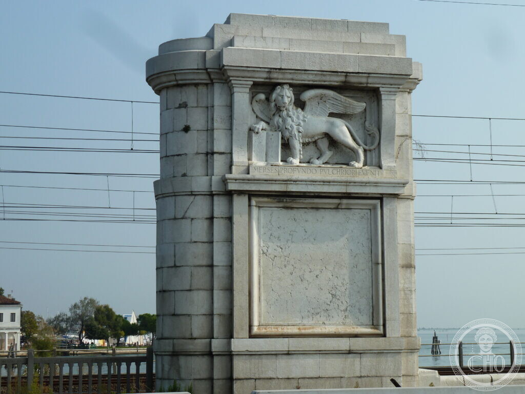 A square stone pier on the Ponte della Libertà carved with the winged lion of Saint Mark, the overhead wires of the railway behind it