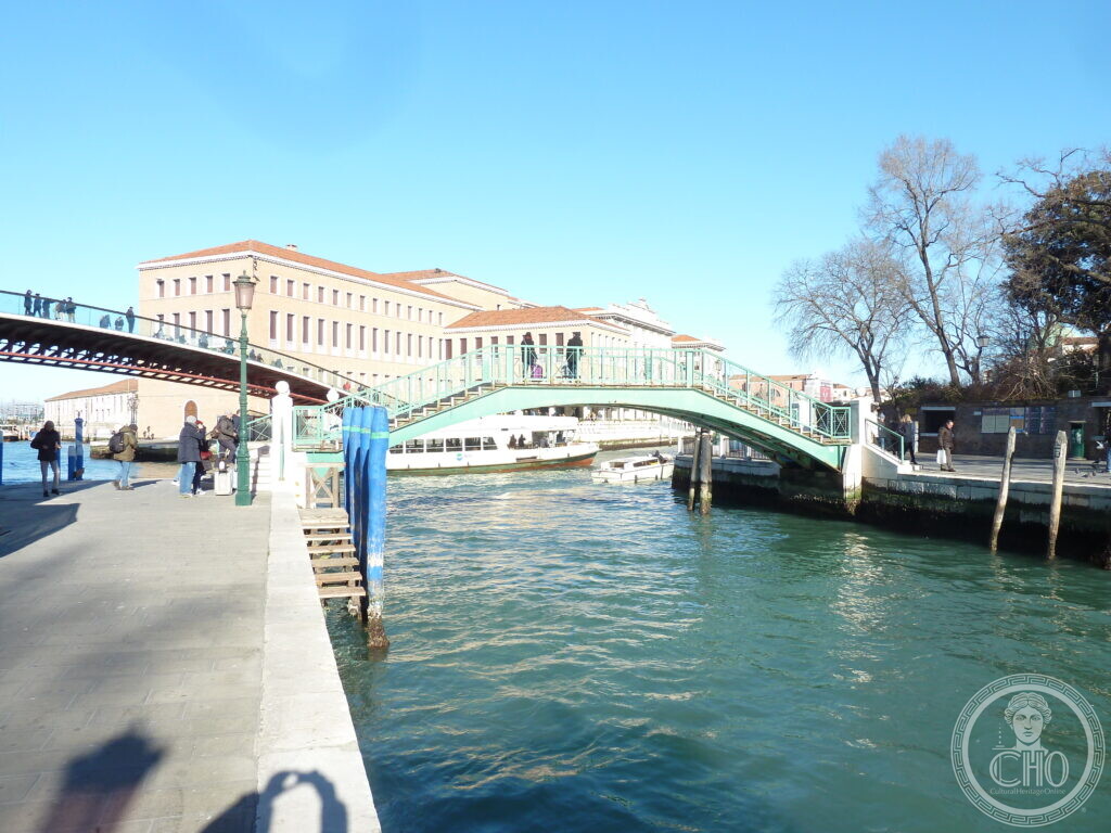 A green-painted iron footbridge arching over a wide canal in Venice, with a long brick and stone building behind it and a second modern bridge to the left