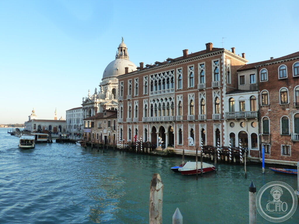 The red-brick neo-Gothic front of Palazzo Genovese on the Grand Canal in Venice, white Gothic loggias on its two upper floors, the dome of the Salute rising behind it at the left and boats on the water