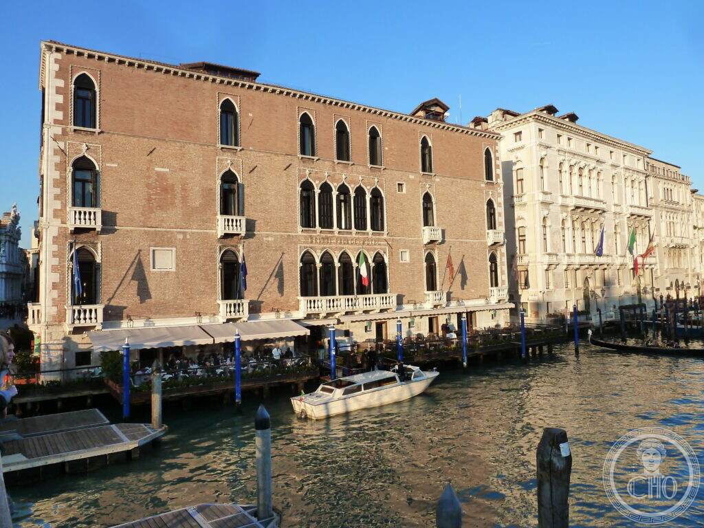 The red-brick Gothic front of Palazzo Pisani Gritti on the Grand Canal in Venice, pointed windows with small balconies, the hotel terrace with its white awnings on the water and a motor launch passing