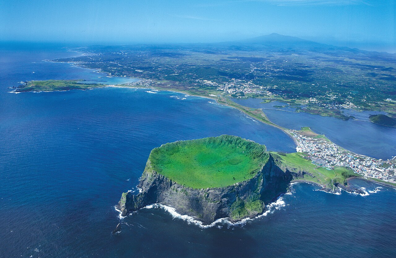 Aerial view of Seongsan Ilchulbong (Sunrise Peak) tuff cone on the eastern tip of Jeju Island, South Korea — a 180-metre-high volcanic tuff cone with a 90,000 square-metre caldera at its summit, rising directly from the sea, inscribed as part of the UNESCO World Heritage Jeju Volcanic Island and Lava Tubes