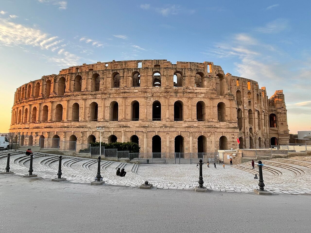 The Roman amphitheatre of El Jem rising from the flat Tunisian steppe, its three-storey elliptical limestone facade intact on three sides, with the gap in the outer wall visible where Turkish forces demolished a section in the 17th century