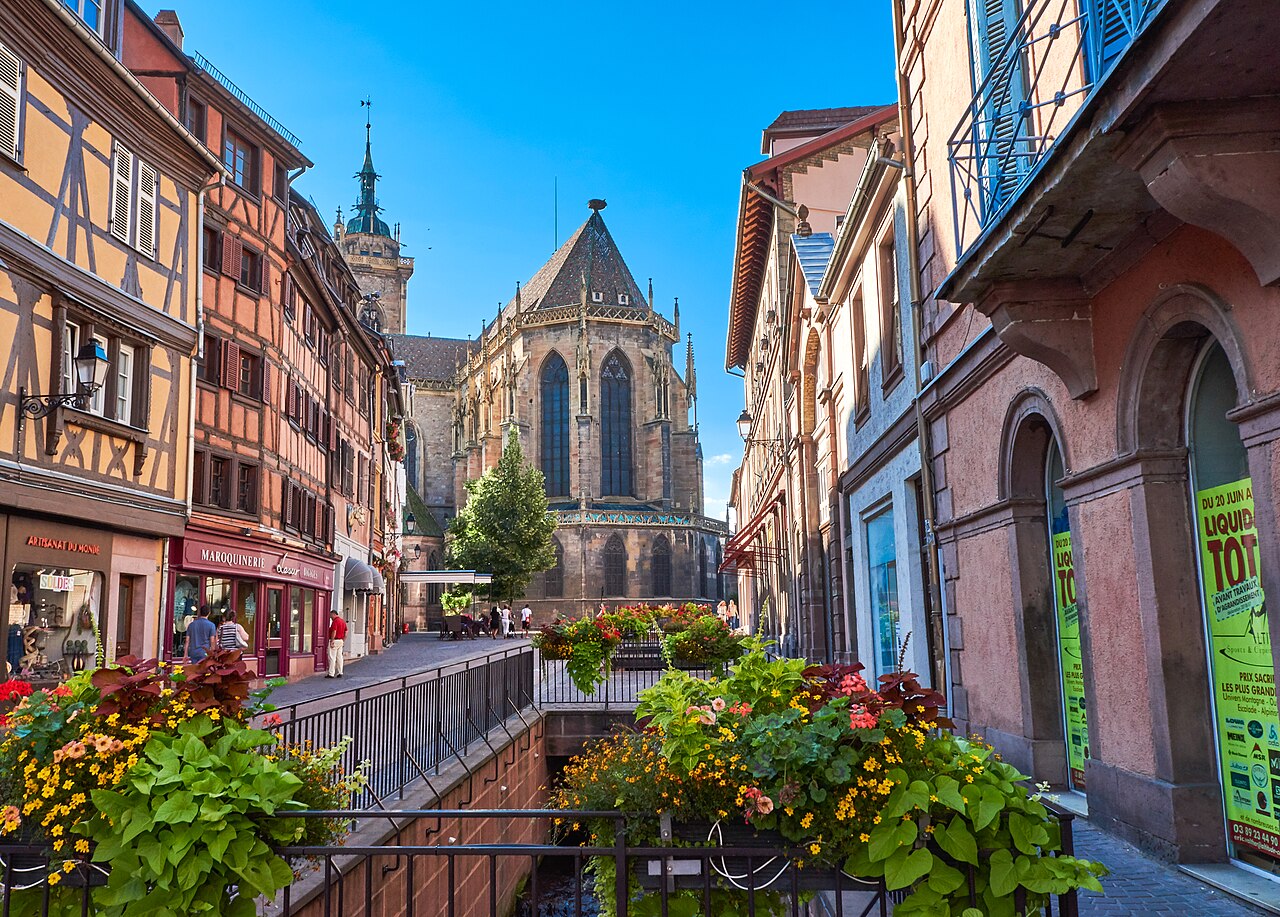 The Gothic collegiate church of Saint-Martin in Colmar, Alsace, its red sandstone tower rising above the medieval half-timbered buildings and canals of the Petite Venise quarter