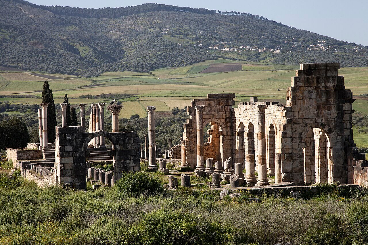 Wide view across the excavated ruins of Volubilis, Morocco, showing the Triumphal Arch of Caracalla and the Zerhoun plain stretching to the horizon