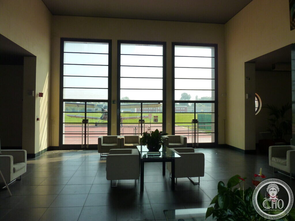 Interior lounge of the Nicelli airport terminal, with large windows looking onto the airfield and a light aircraft parked outside