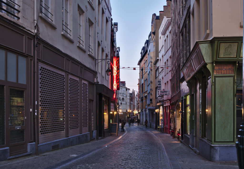 Narrow cobbled Rue des Éperonniers in Brussels at dusk, seen from Place Saint-Jean, with yellow brick fronts on the right carrying the vertical lettering hotel alma