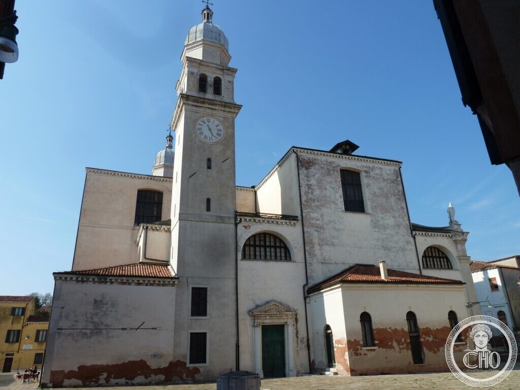 A pale plastered church with a tall square bell tower carrying a clock and a lead-covered cupola, seen from a paved square