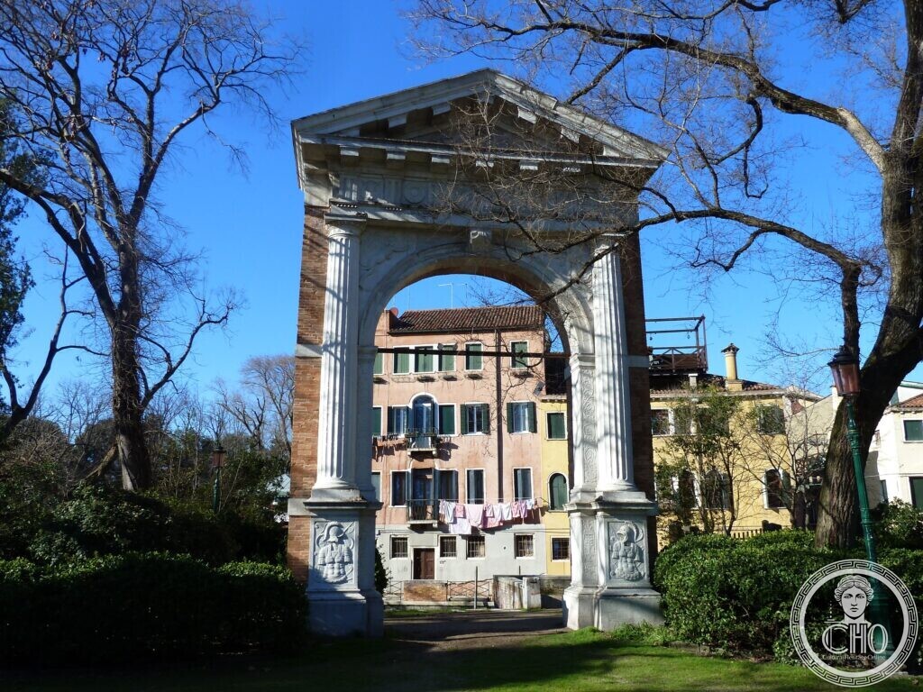 A classical stone archway with fluted columns and a triangular pediment standing alone on a lawn among bare trees, houses visible through it