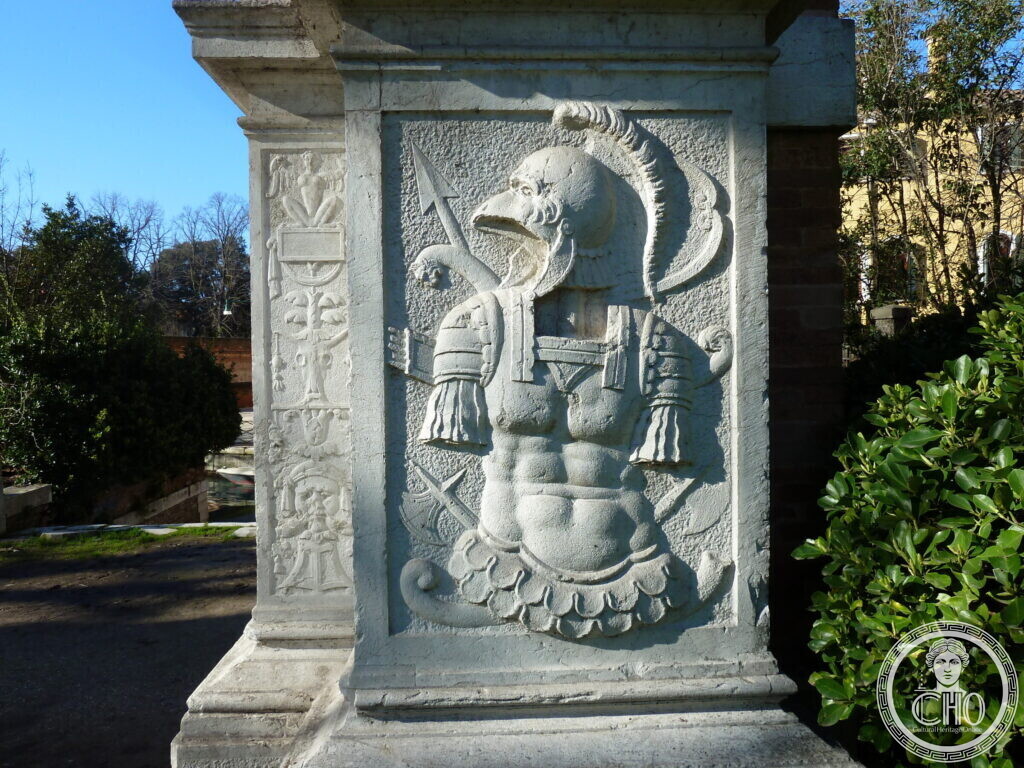 A white stone pedestal carved in relief with a helmeted grotesque head over a trophy of armour and weapons