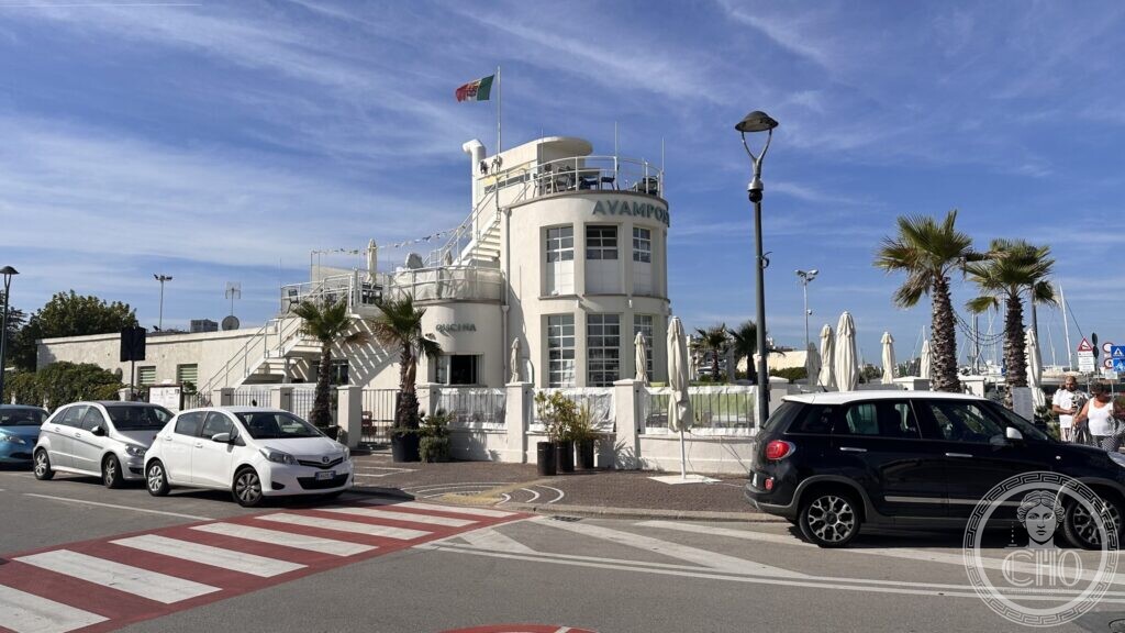 The white building seen from the quayside road, with parked cars, palms and the flag on the turret
