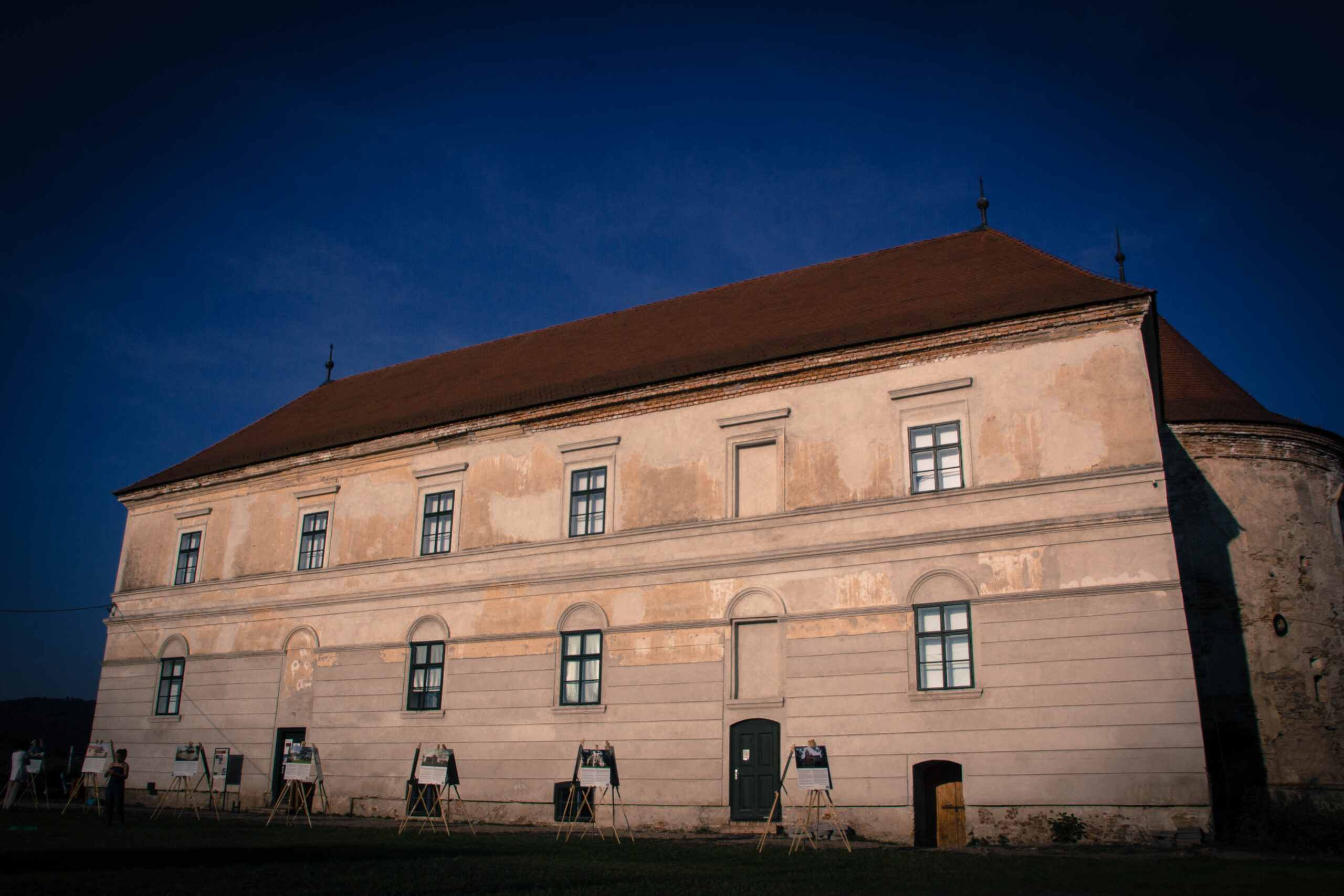 The restored main range of the Bánffy castle at Bonțida at dusk, with an outdoor photographic exhibition in front of it
