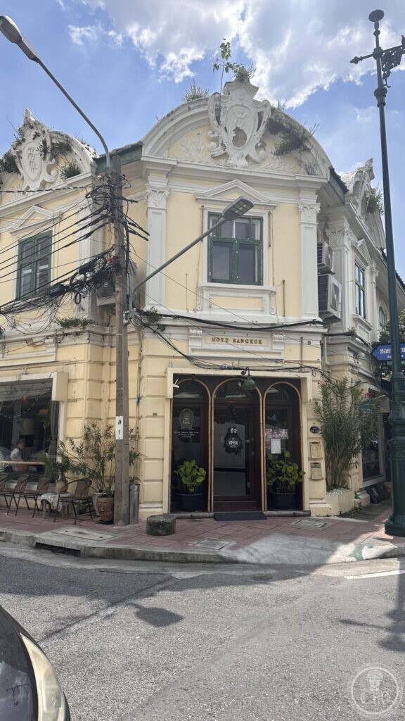 A pale yellow corner building with a curved pediment, carved cartouche and green-shuttered windows on Soi Phraya Si, with plants growing from the cornice