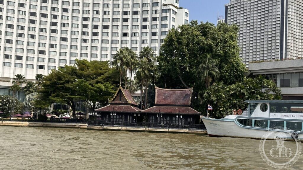 A dark teak pavilion with a tiered tiled roof standing on the bank of the Chao Phraya directly in front of two large modern hotel blocks