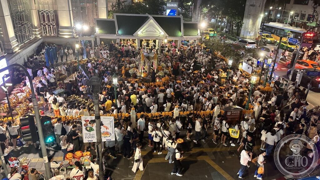 A dense crowd around the Erawan Shrine at the Ratchaprasong intersection in Bangkok at night, with marigold garlands, shopping malls behind and two food-campaign banners hanging on the railing