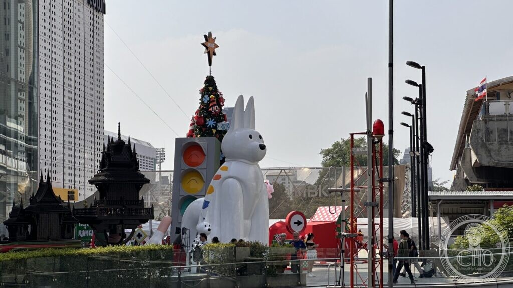A giant white rabbit sculpture and a Christmas tree installed on a plaza outside a Bangkok shopping centre, next to a dark carved model of a traditional Thai pavilion