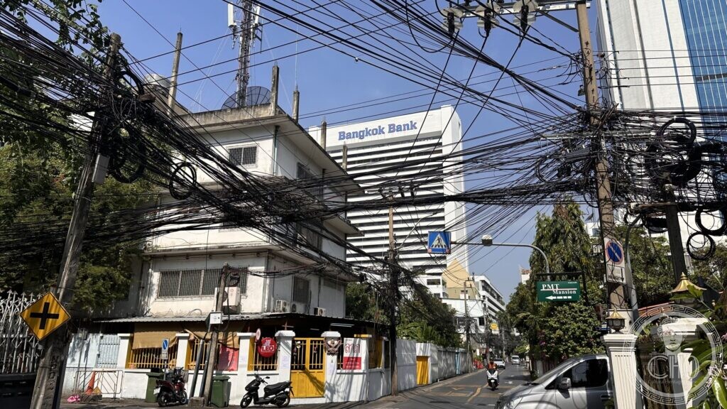 A tangle of overhead electrical cables above a Bangkok street corner, with a small yellow-gated shrine building below and a bank tower behind