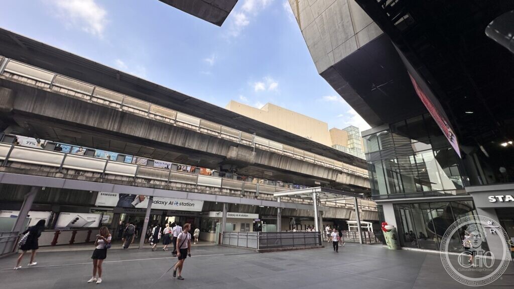 The concrete underside of the elevated railway at a Bangkok interchange, with advertising panels and a station entrance branded as a phone-maker's AI station