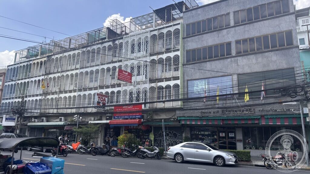 A long four-storey row of shophouses on Ti Thong Road, at the corner of Ratchabophit Road, with arched screen openings on every floor, shopfronts below and motorbikes parked outside