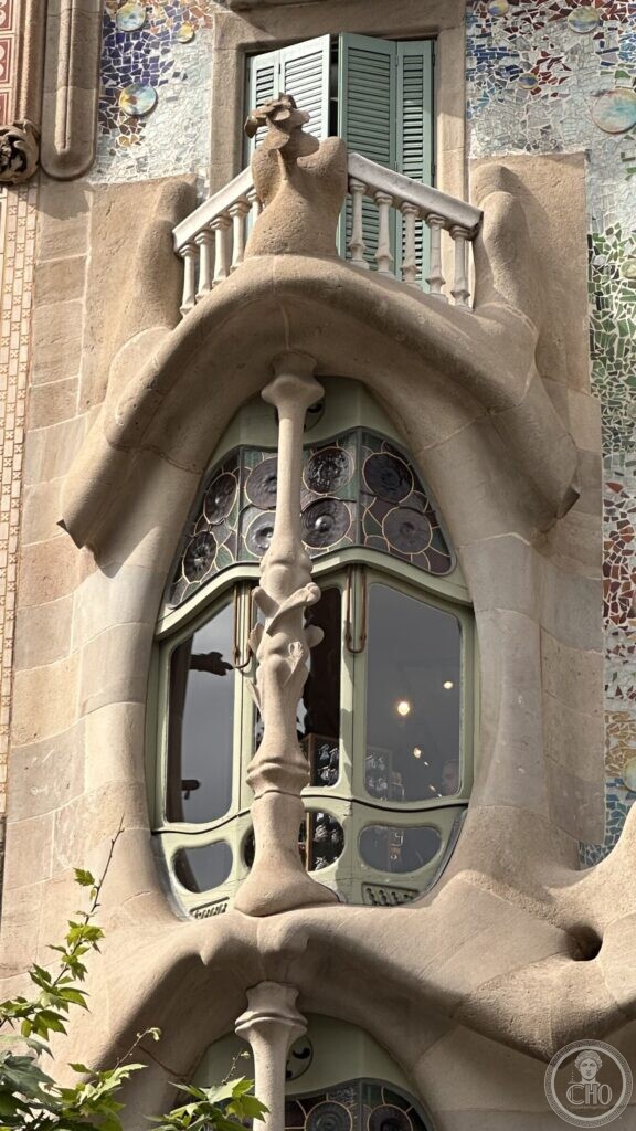 A bone-like stone window frame with stained glass and a twisted colonnette, set in a wall of coloured mosaic
