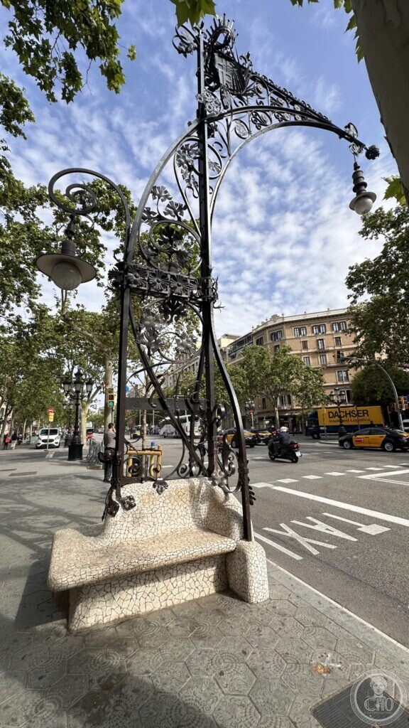A wrought-iron street lamp curving over a white mosaic bench, on a wide pavement