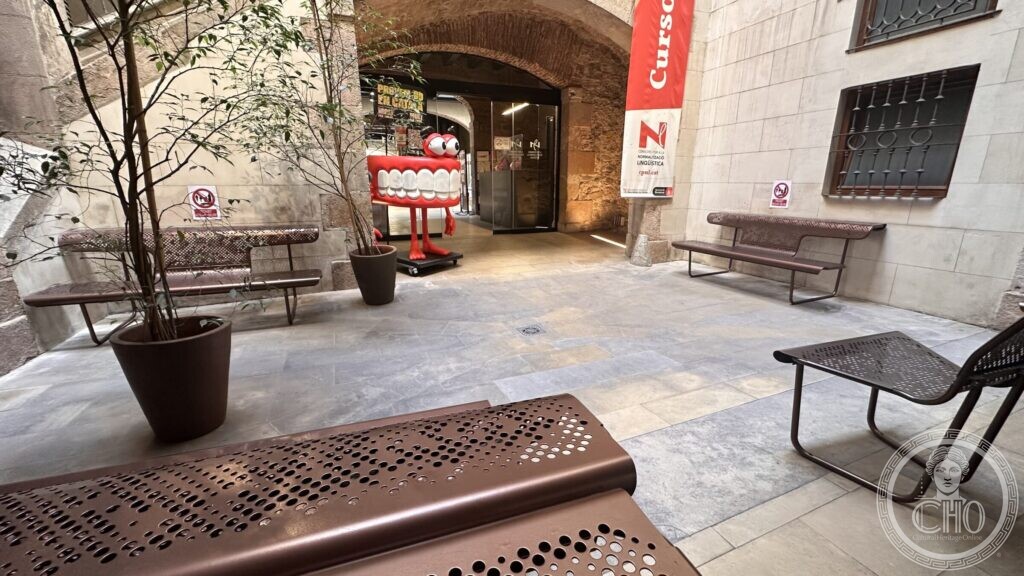 A small courtyard with benches, potted trees and a red sculpture under a brick vault