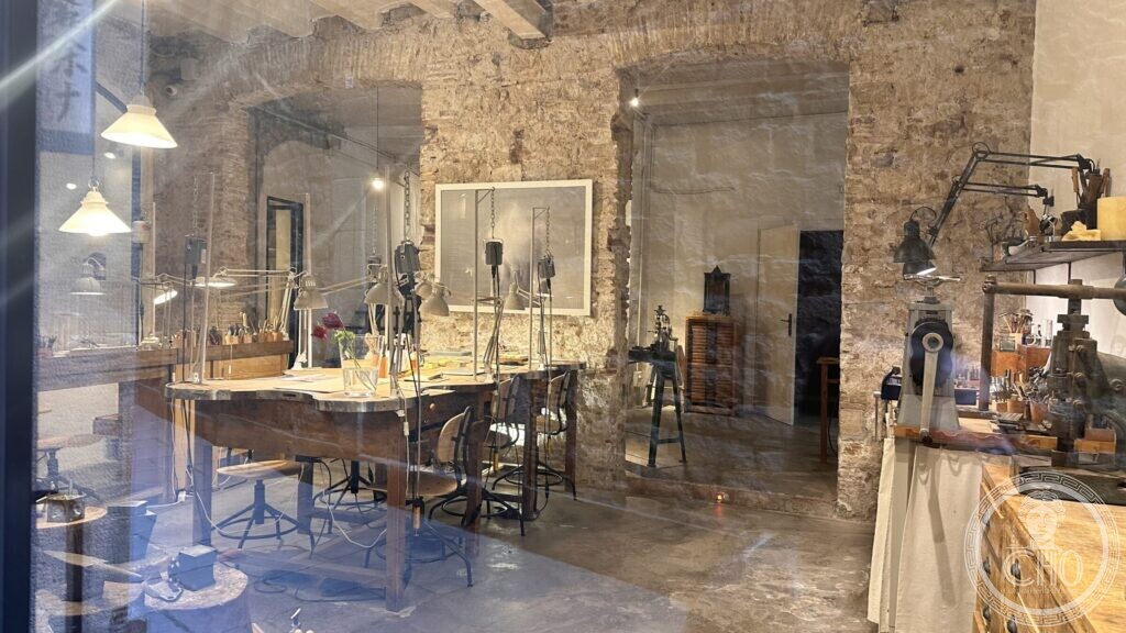 A goldsmith’s workbench seen through a shop window, with lamps on arms and a bare stone wall behind