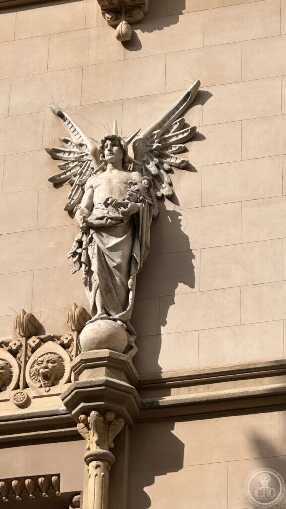 A winged stone figure holding a bundle, standing on a globe on a corbel high on a façade