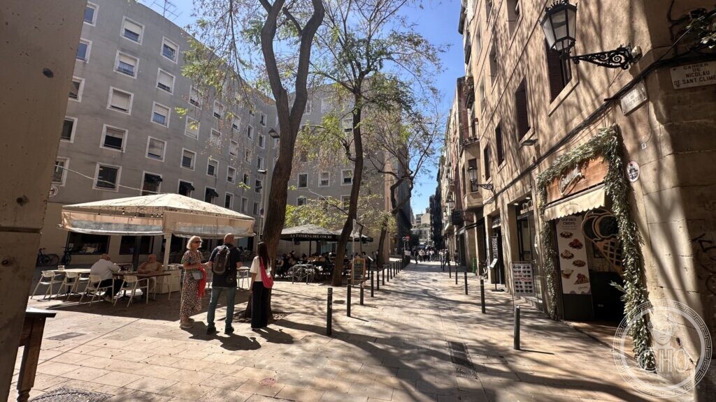 A quiet paved street in La Ribera with a café awning, plane trees and residents walking