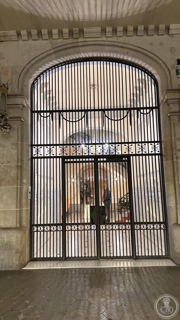 A wide iron gate under a shallow arch, with a chandelier visible in the hall beyond
