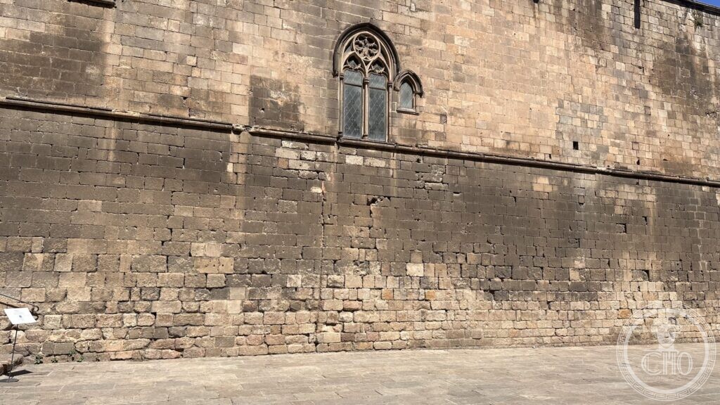 A high ashlar wall with a single Gothic window, above a paved square