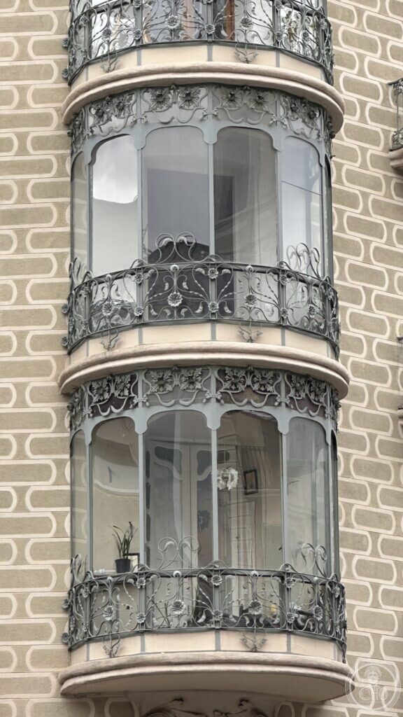 A rounded glazed bay window with iron railings of flowers, against a wall of scrolled sgraffito