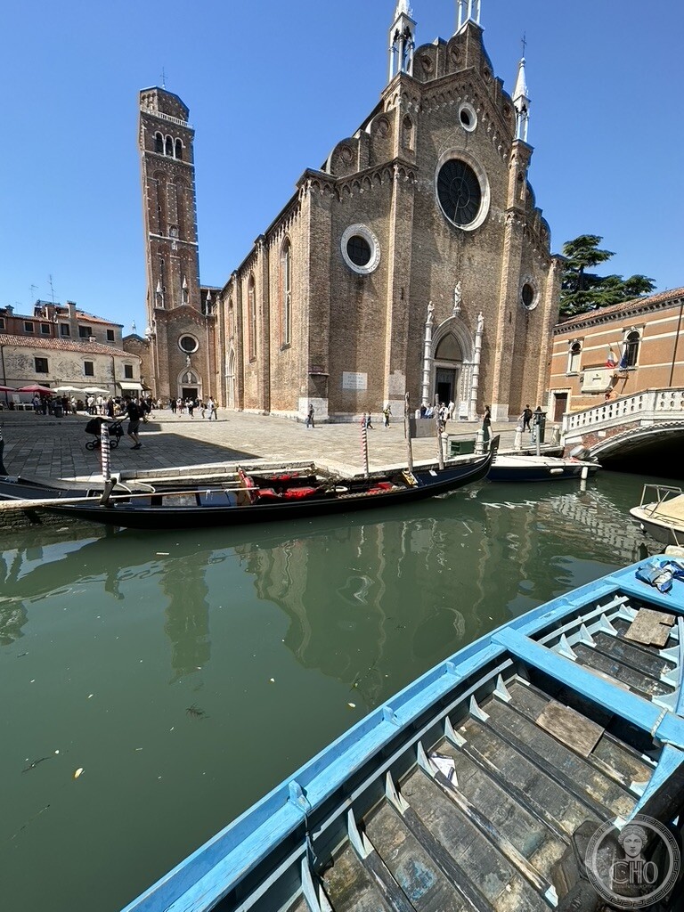 The brick front and campanile of the Frari seen across the Rio dei Frari, a gondola moored along the fondamenta and the bow of a blue boat in the foreground