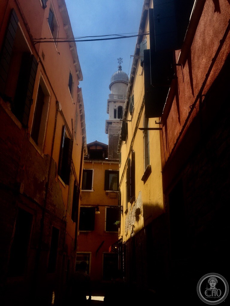A narrow calle beside the Scuola in shadow, a bell tower with an onion dome rising against the sky at the end of it