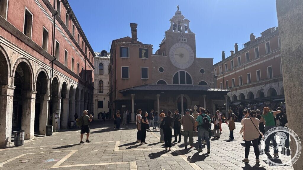 A group of people standing in a paved square under arcades, facing a small brick church with a large clock and bell gable
