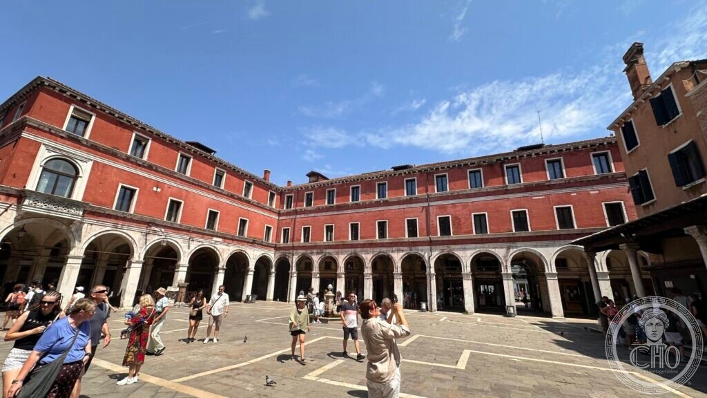 The open paving of Campo San Giacomo di Rialto with visitors crossing it, arcaded buildings in red plaster on two sides