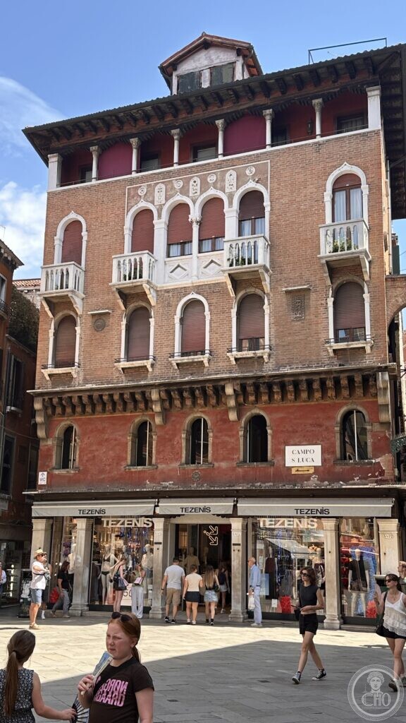 Full facade of a Gothic-Venetian palace on Campo San Luca, with a rooftop timber altana and Tezenis shopfront at street level