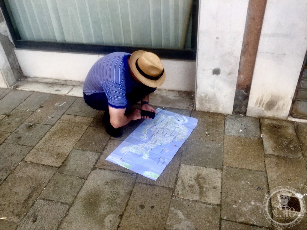 A traveler wearing a fedora crouched on the stone pavement reading an unfolded paper map of Venice