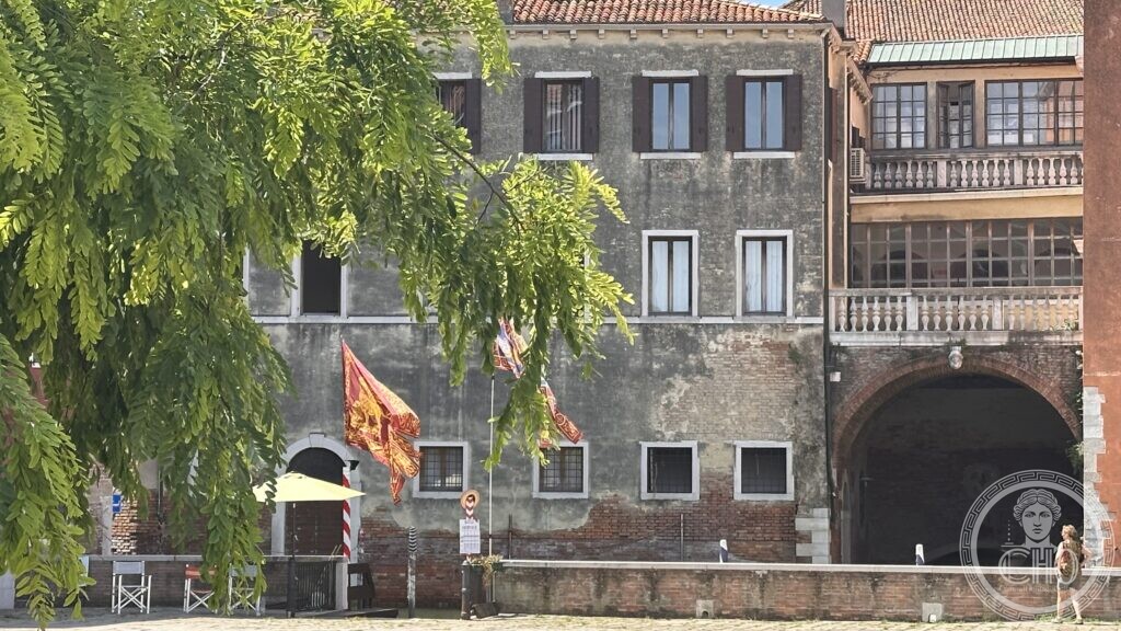 A weathered Venetian palace with Venetian flags flying, seen across Campo Sant'Angelo through tree branches, with an archway leading to the Rio di Sant'Angelo
