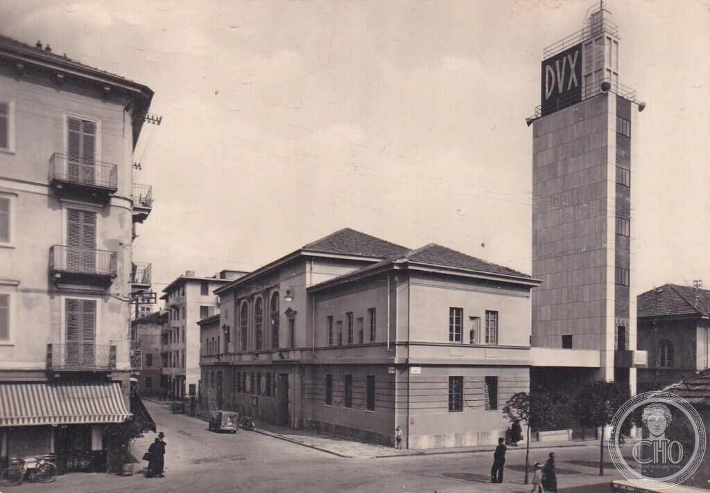 A period postcard of the Torre Littoria in Biella, a tall pale tower with the letters DVX at the top rising at a street corner beside a two-storey building, passers-by in the street