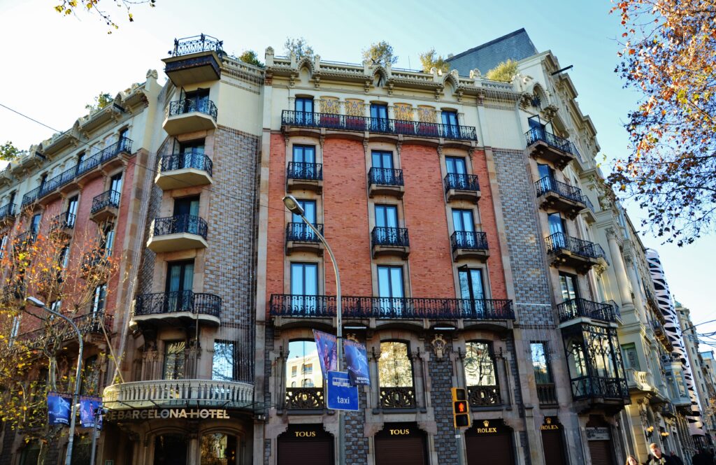 Corner building in red exposed brick framed by bands of grey and brown stone, with wrought-iron balconies and a polygonal corner body, Passeig de Gràcia 75, Barcelona