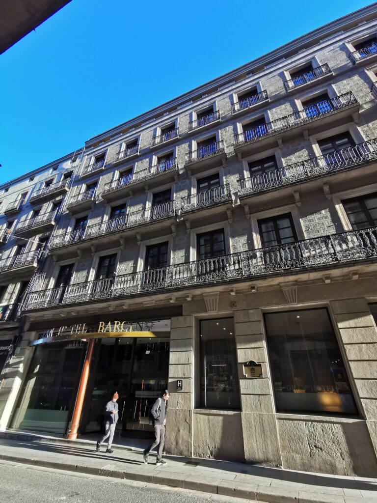Long stone facade with a continuous wrought-iron balcony on the first floor and individual balconies above, hotel entrance at street level, Carrer de Jaume I 6, Barcelona