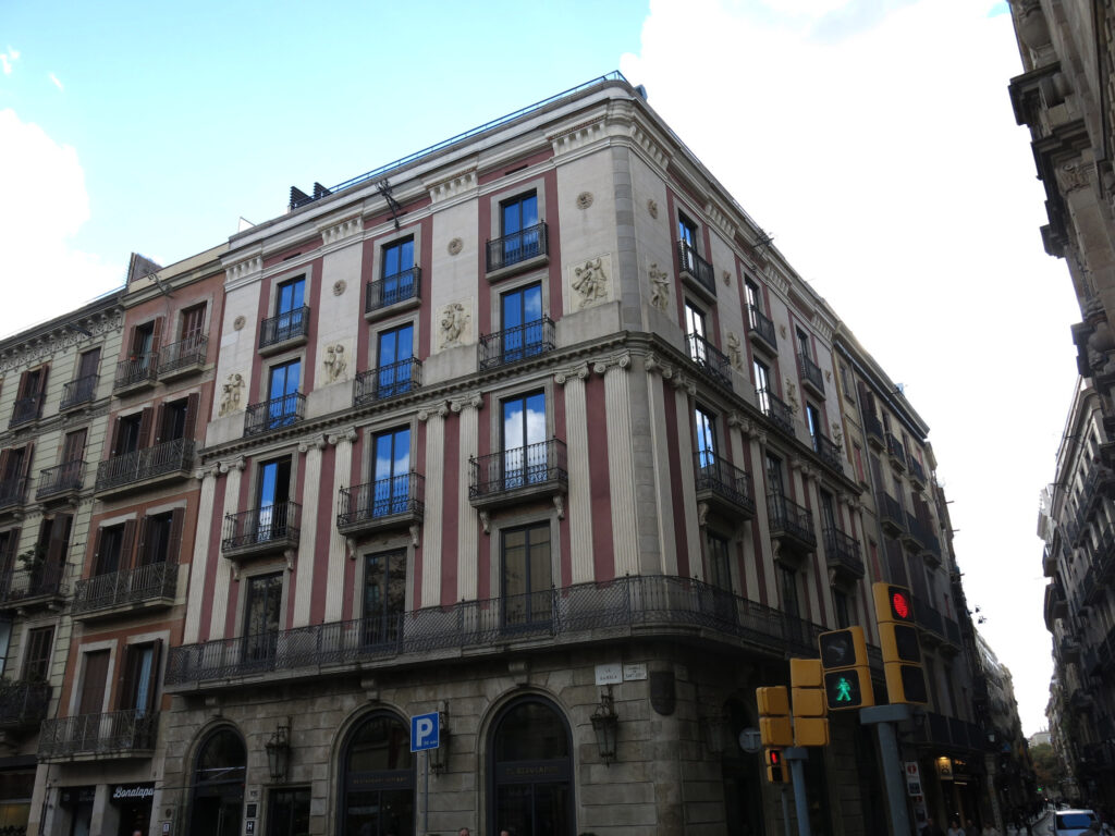 Corner building with pink stucco, paired white pilasters, terracotta reliefs of children and a stone ground floor with tall round arches, La Rambla 105, Barcelona