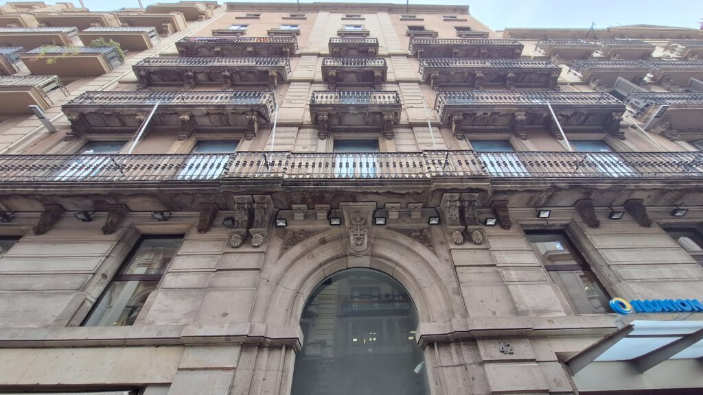 Stone ground floor with a large round-arched central portal, continuous iron balcony and paired balconies on carved brackets above, Via Laietana 42, Barcelona