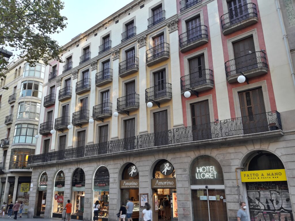 Long facade on La Rambla with a continuous wrought-iron balcony, pilasters with Corinthian capitals, cream and pink bays and shops in round arches, Rambla 125, Barcelona