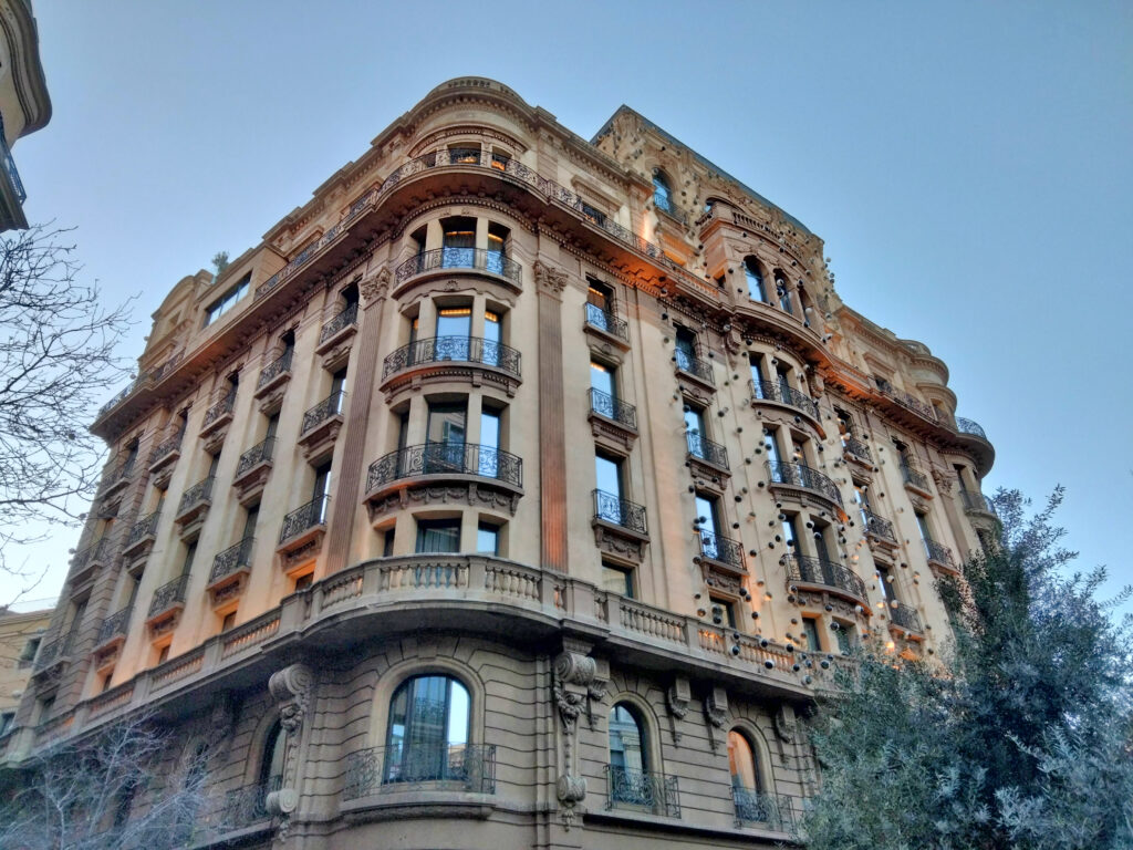 Rounded corner building in stone-coloured stucco with curved balconies, a balustrade and scattered small ceramic eyes on the facade, Via Laietana 49, Barcelona