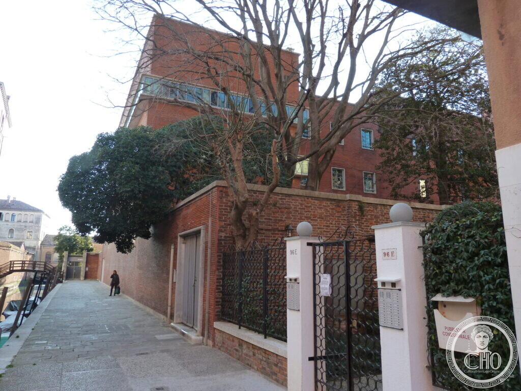 A brick building with ribbon windows rising behind a garden wall, gate and trees along a canal-side walkway
