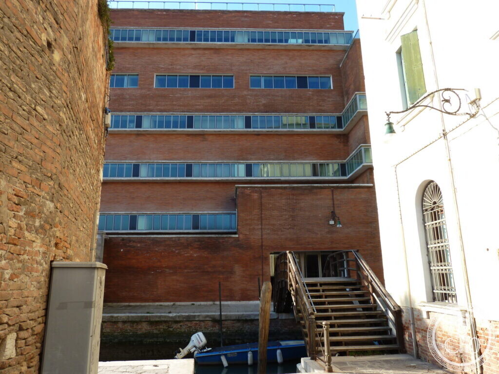 A brick industrial building with long horizontal bands of windows rising above a canal, a wooden stair down to the water