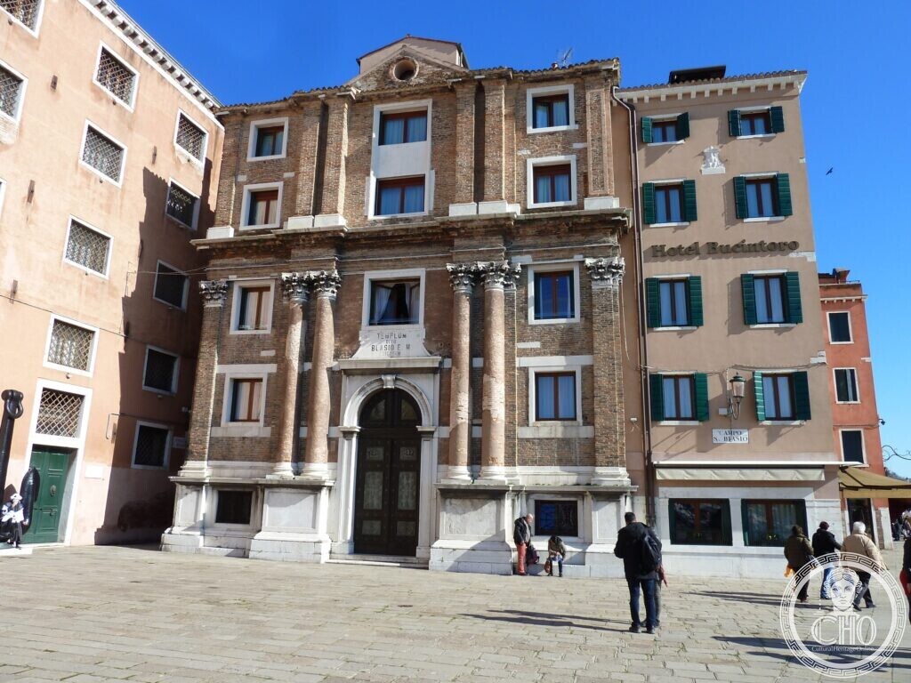 A church front in red brick and white stone with two orders of paired columns and a Latin inscription over the door, between two plastered buildings
